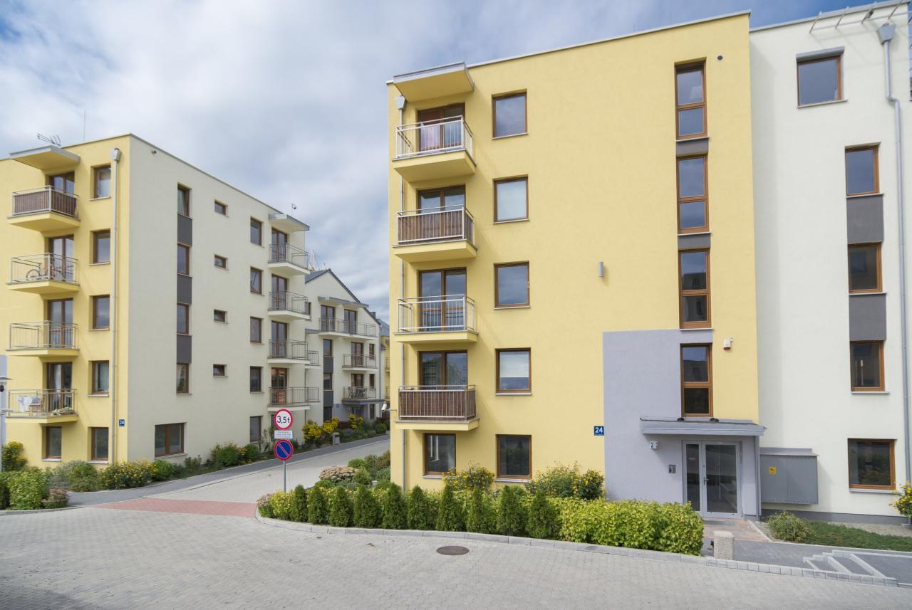 Nowoczesne budynki wielorodzinne w pastelowych kolorach z balkonami, otoczone zielenią. Jeden z budynków ma żółtą elewację.