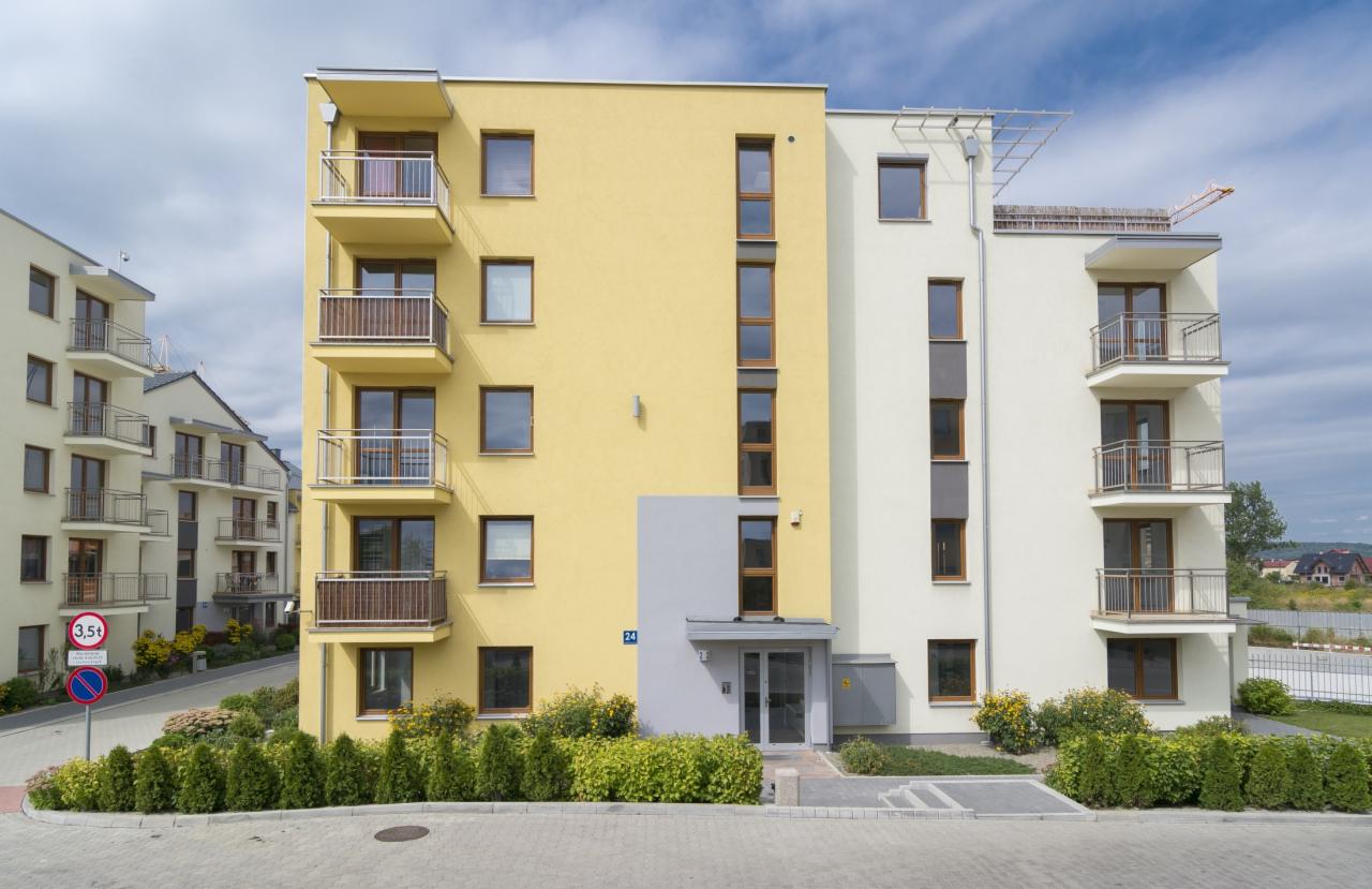 Nowoczesny, wielopiętrowy budynek mieszkalny z balkonami, o elewacji w odcieniach żółci i bieli, z subtelnymi szarymi akcentami. Widok z poziomu chodnika, zieleń w tle.