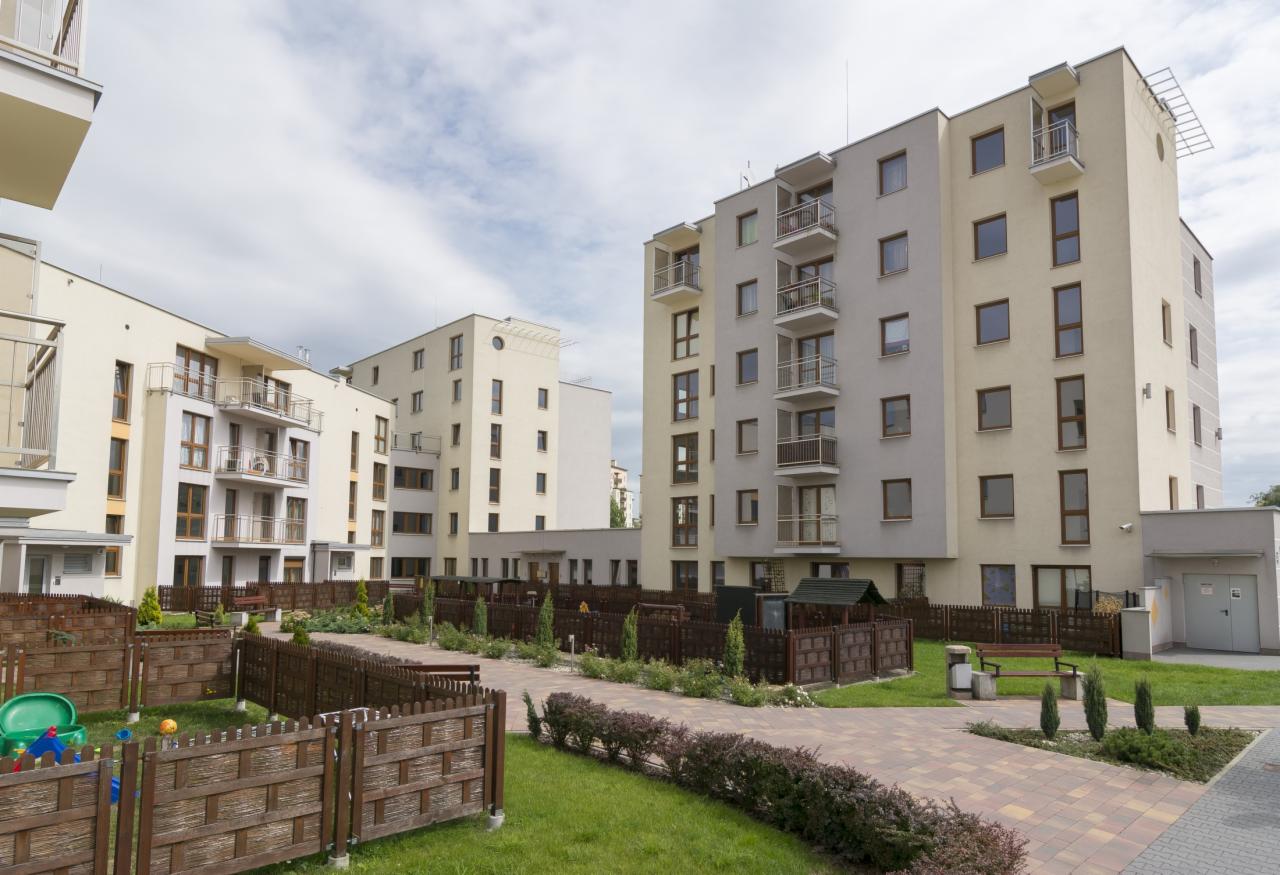 Nowoczesne budynki wielorodzinne z balkonami, oddzielone niskim, drewnianym ogrodzeniem i zadbaną zielenią, tworzące estetyczne osiedle mieszkaniowe.