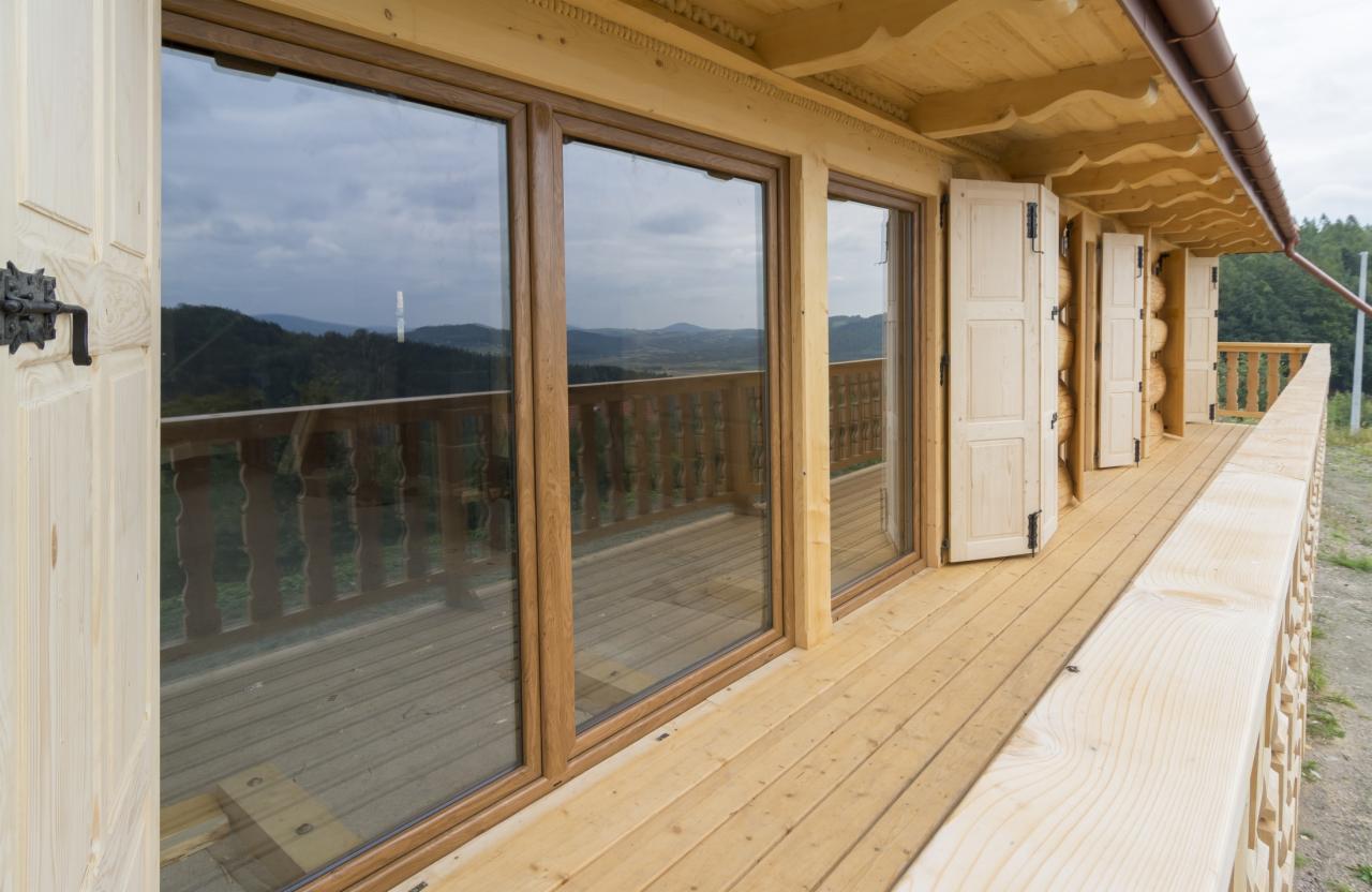 Drewniany balkon z balustradą i trzema oknami z widokiem na góry. Otwarta drewniana furtka z ozdobną klamką. Otwierane okiennice.