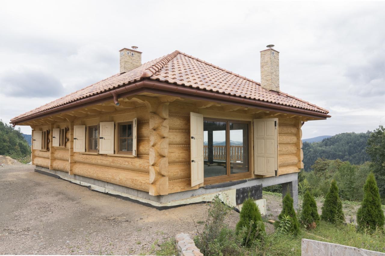 Drewniany dom z bali, wykończony czerwoną dachówką, z otwartymi okiennicami i widokiem na góry. Budynek na lekkim podwyższeniu, z fragmentami izolacji widocznymi u podstawy.