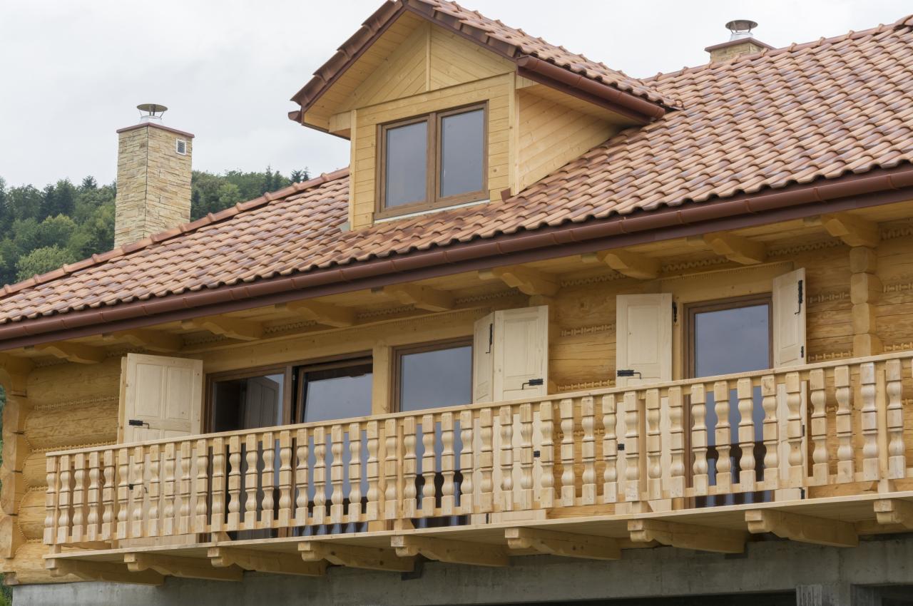 Drewniany dom z balkonem i ceramiczną dachówką. Detale zdobień na elewacji, otwarte okiennice. Kompozycja z zielenią w tle.