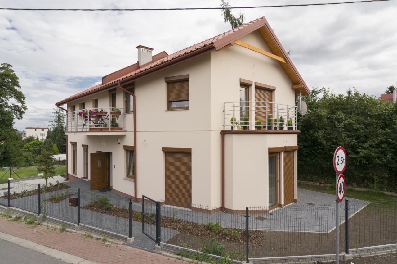 Dwupiętrowy dom z beżową elewacją, brązowymi roletami i czerwonym dachem, widziany z poziomu ulicy. Otwarta brama i drzwi wejściowe.