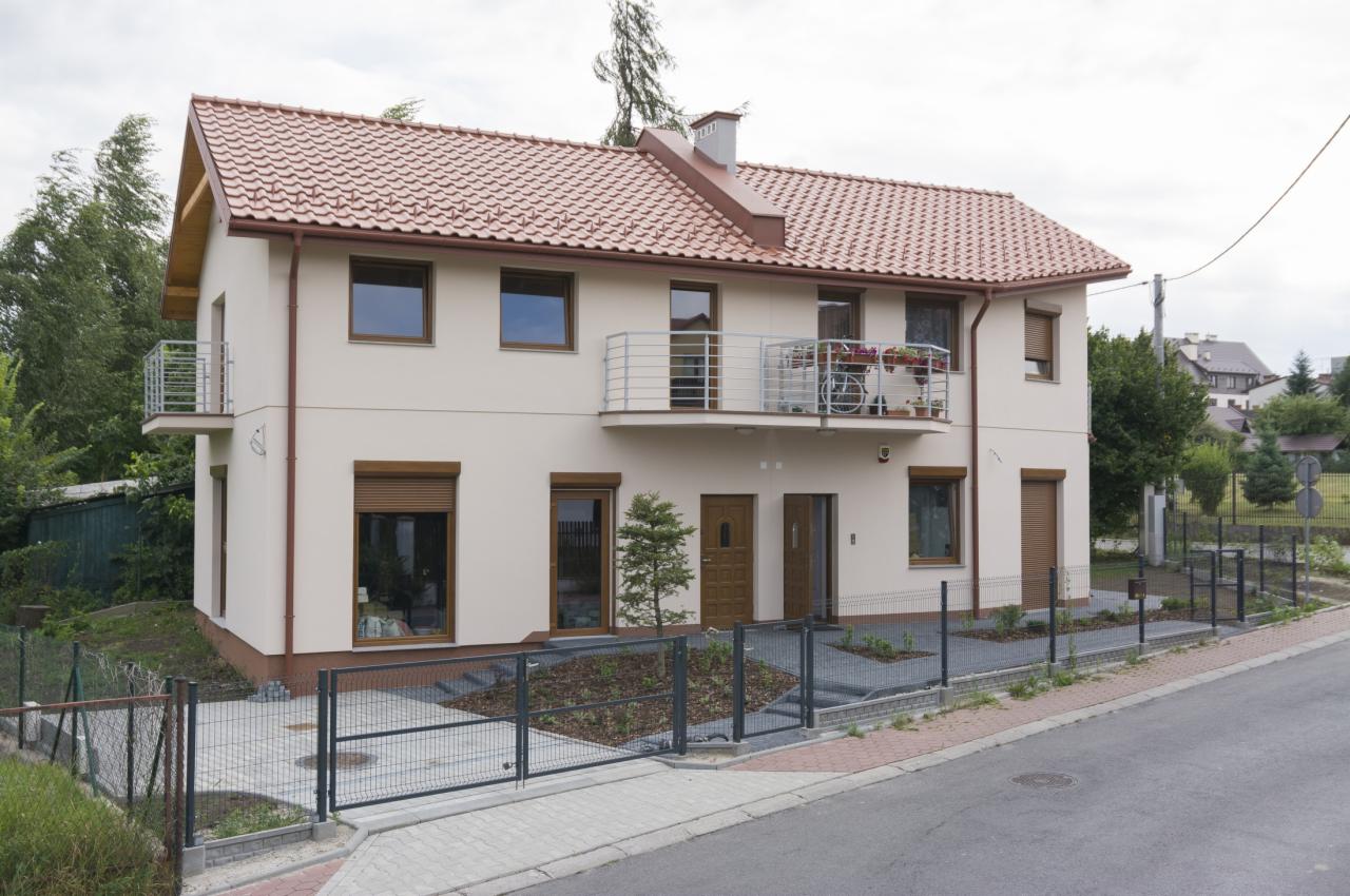 Dwupiętrowy dom z jasną elewacją, balkonem z metalową barierką i rowerem, brązowymi oknami i drzwiami, oraz ogrodzeniem z siatki. Widok z ulicy.