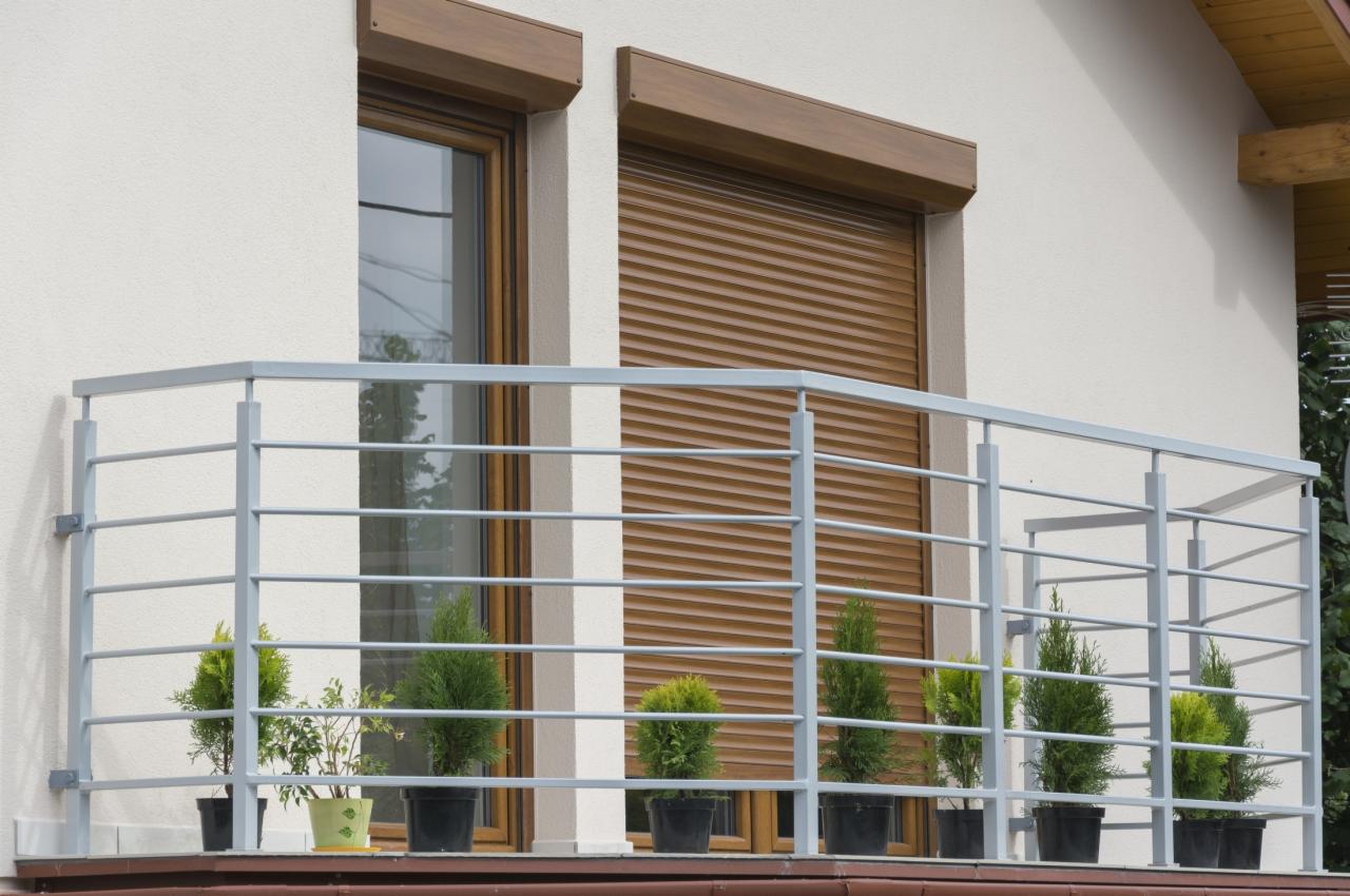 Szary balkon z roślinami w doniczkach na tle brązowych rolet i okna. Elementy drewniane w górnej części okien. Jasna elewacja budynku.
