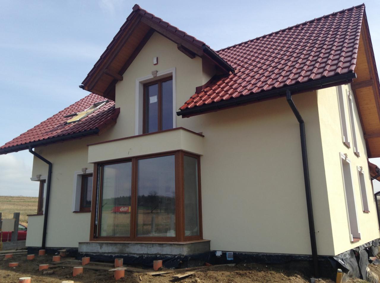 Elewacja domu jednorodzinnego z czerwoną dachówką i dużymi oknami, widok z boku w słoneczny dzień. Widoczne elementy wykończeniowe i teren wokół domu w trakcie prac.