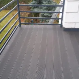 Nowoczesny balkon z szarą, kompozytową podłogą, metalową balustradą i widokiem na zieleń. Drabina oparta o ścianę w tle.