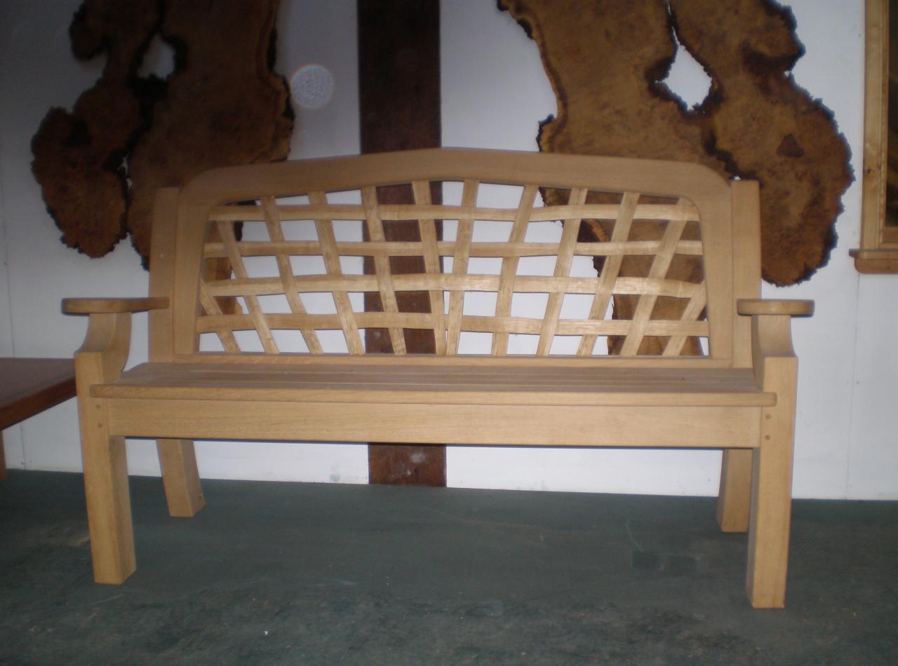 Drewniana ławka ogrodowa z ażurowym oparciem, widoczna na tle ściany z dekoracjami z drewna. Ławka w jasnym odcieniu drewna, o prostej, klasycznej formie.