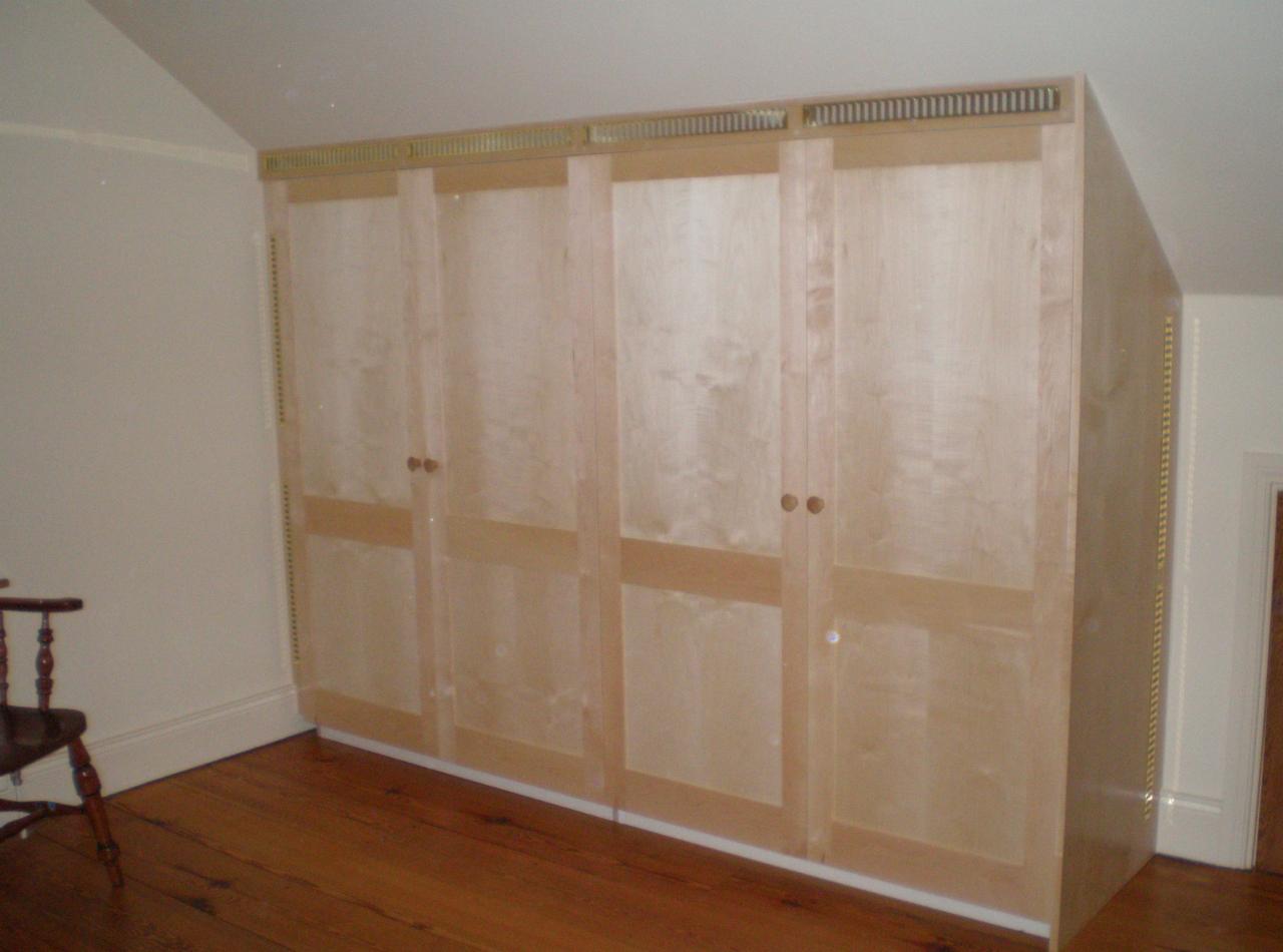 Czterodrzwiowa szafa z jasnego drewna, wbudowana w skos pomieszczenia na poddaszu, z widoczną wentylacją u góry i drewnianą podłogą.