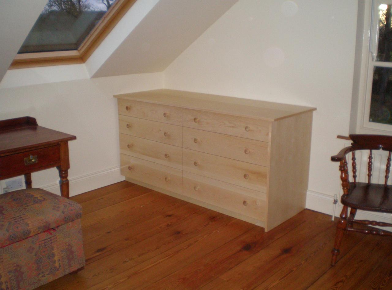 Jasna komoda z drewna z ośmioma szufladami, dopasowana do skosu poddasza. Obok biurko, krzesło i okno. Drewniana podłoga.