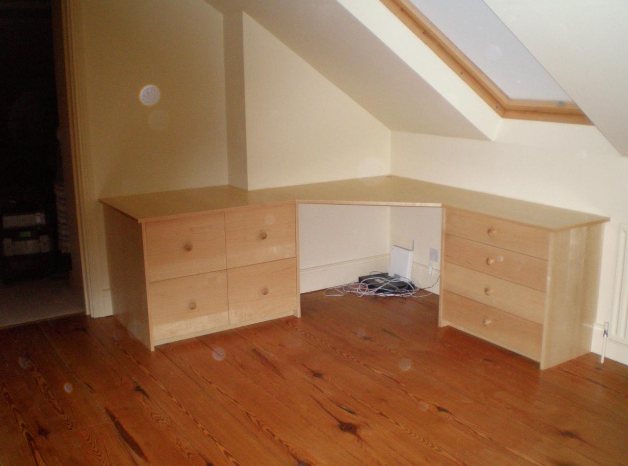 Jasne drewniane szafki z szufladami i blatem w rogu pokoju na poddaszu z oknem dachowym, tworzące przestrzeń do pracy. Widoczna drewniana podłoga.