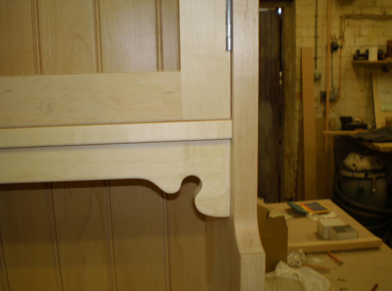 Fragment jasnej, drewnianej szafki z frezowanym detalem pod blatem. W tle warsztat stolarski z narzędziami.