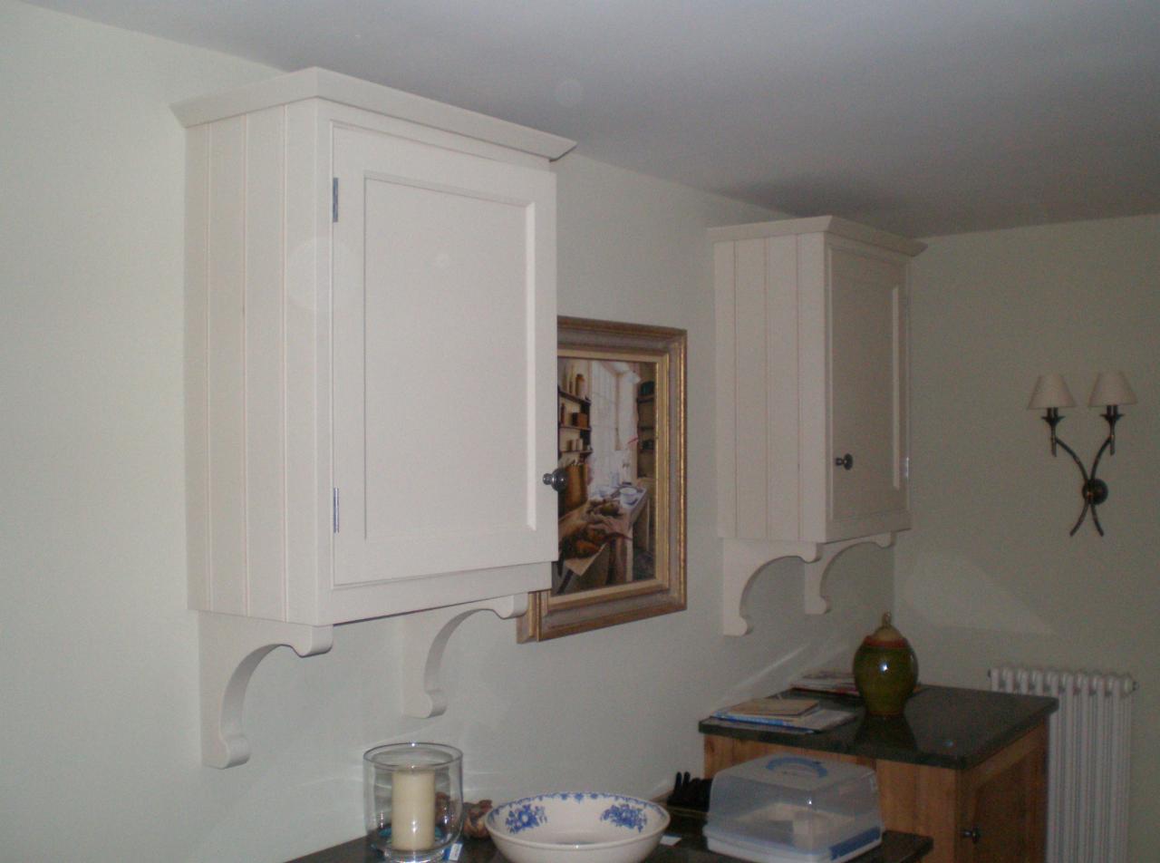 Dwie białe, wiszące szafki kuchenne z dekoracyjnymi wspornikami, jedna z lekko otwartymi drzwiczkami, na tle ściany z obrazem i kinkietem.