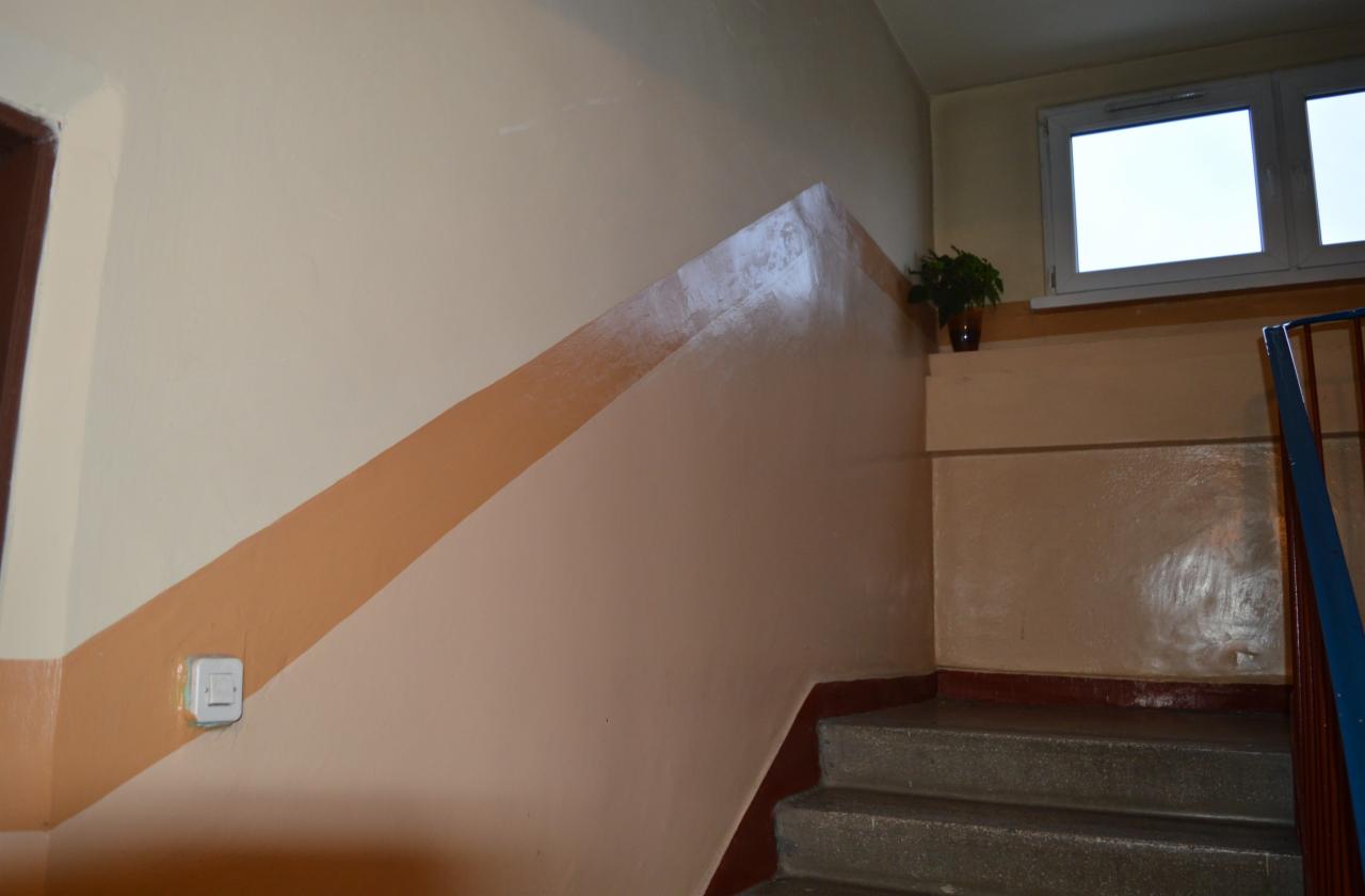Klatka schodowa po malowaniu z beżowymi ścianami i brązowym pasem wzdłuż schodów. Na podeście doniczka z rośliną. Widoczne schody i okno.
