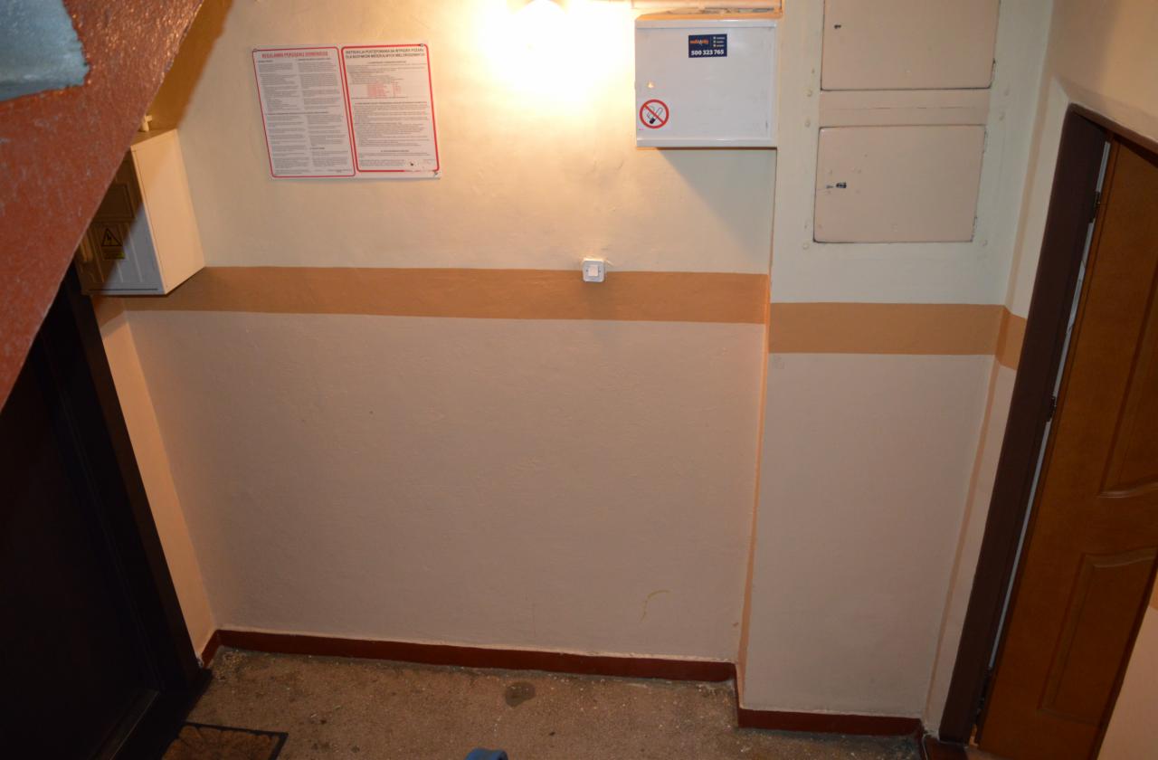 Klatka schodowa po malowaniu z beżowymi pasami, widoczne tablice informacyjne i skrzynka elektryczna. Ujęcie z dołu, lekko z prawej strony.