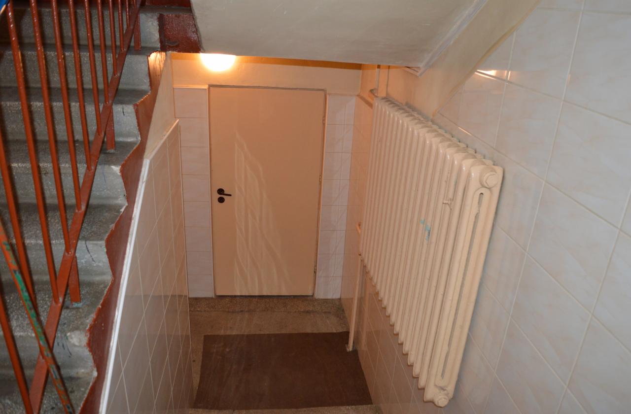 Wnętrze klatki schodowej z beżową, kwadratową glazurą, grzejnikiem żeberkowym i pomalowaną na beżowo futryną drzwi. Widoczne także metalowe schody.