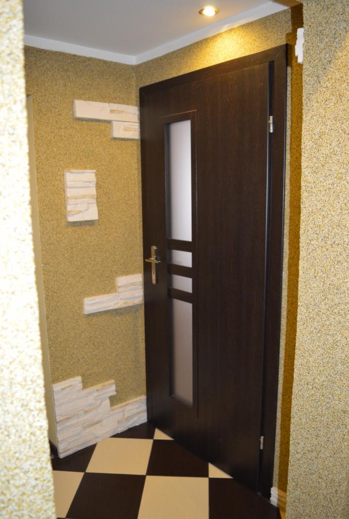 Ciemne drzwi z szybą w otworze. Dekoracja ściany kamieniem dekoracyjnym. Podłoga w szachownicę. Ściany pokryte tynkiem strukturalnym. Ciepłe oświetlenie.