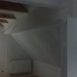 Poddasze po remoncie: białe ściany, drewniane belki stropowe, okno z widokiem na zewnątrz, grzejnik. Wnętrze jest jasne i przestronne, widać elementy wykończeniowe.