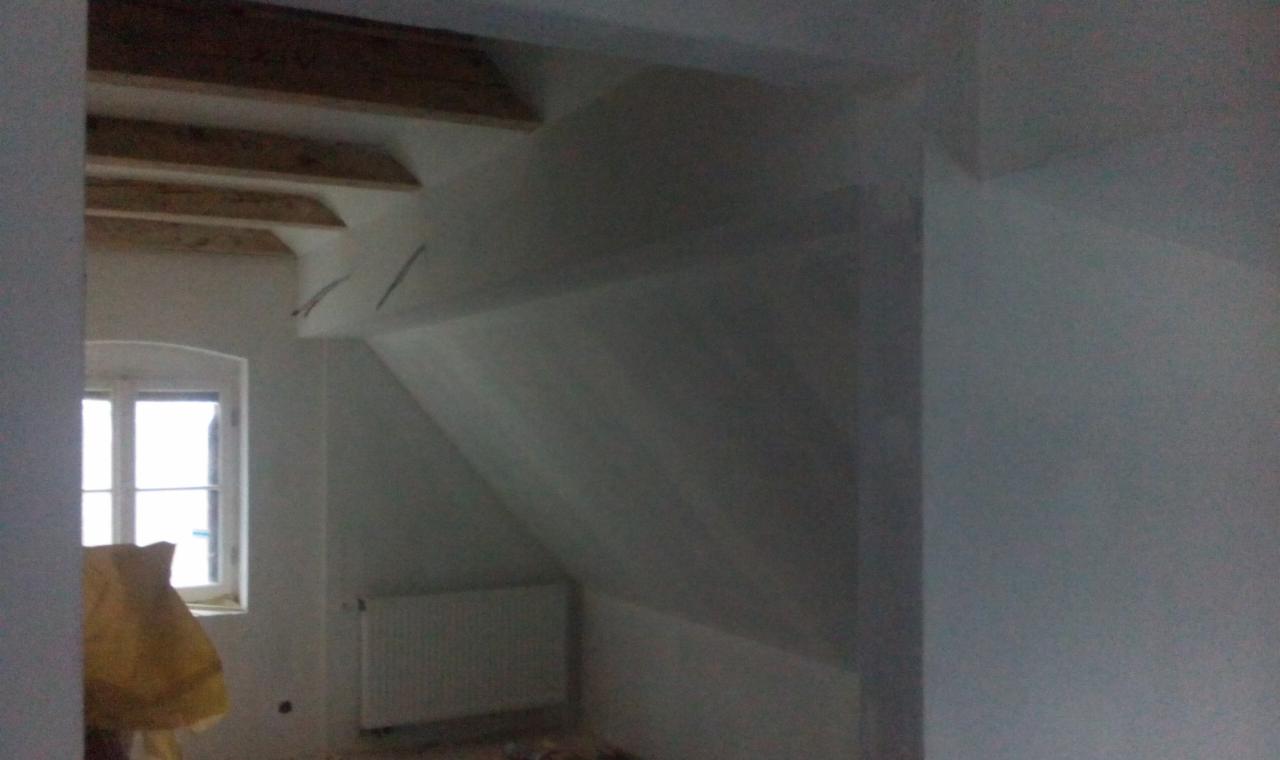 Poddasze po remoncie: białe ściany, drewniane belki stropowe, okno z widokiem na zewnątrz, grzejnik. Wnętrze jest jasne i przestronne, widać elementy wykończeniowe.