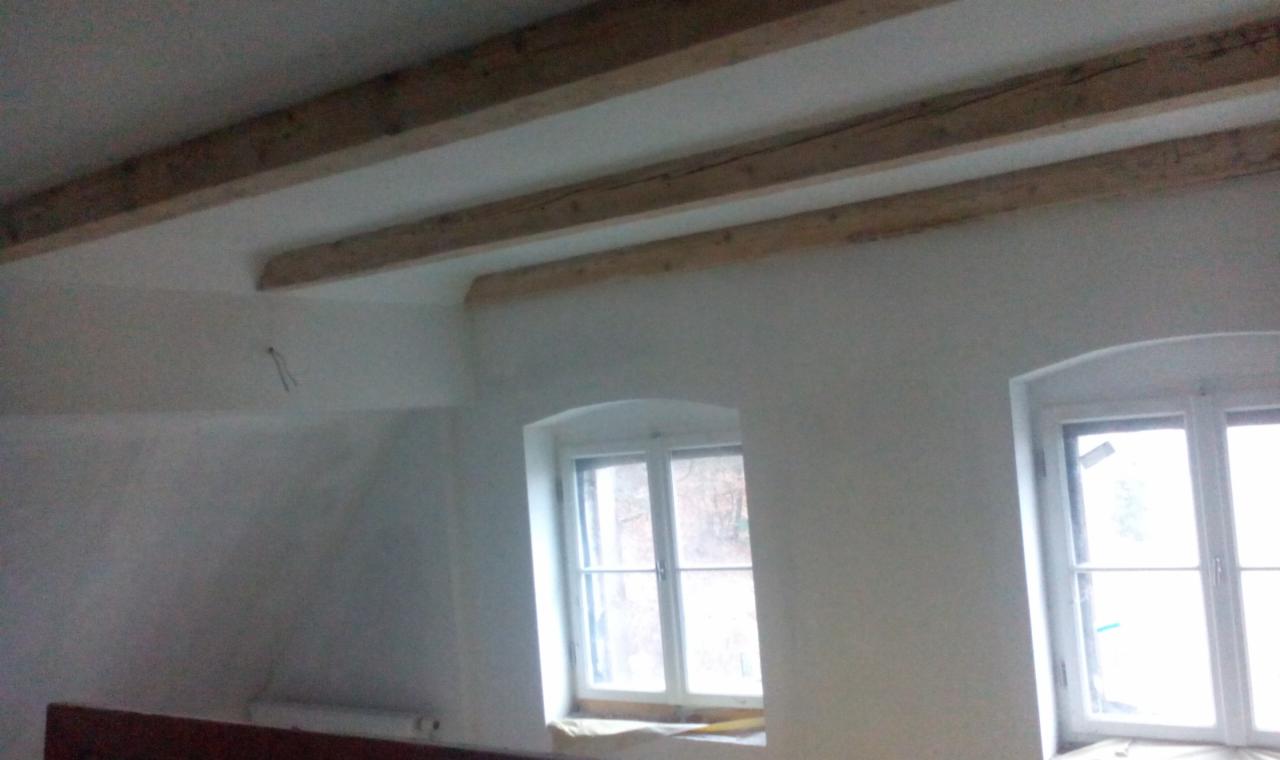 Jasne wnętrze z drewnianymi belkami stropowymi i dwoma oknami w białych ramach. Ściany świeżo otynkowane, fragment instalacji elektrycznej widoczny na ścianie.