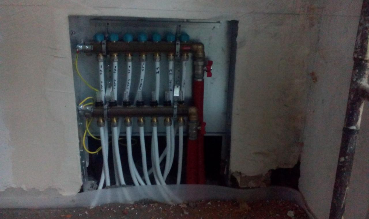 Rozdzielacz ogrzewania podłogowego z niebieskimi pokrętłami, zamontowany w surowej wnęce ściany, z widocznymi rurami i instalacją elektryczną.
