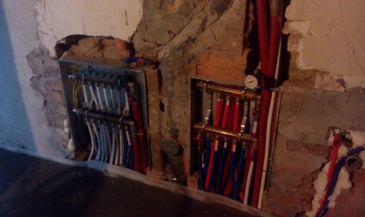 Rozdzielacze instalacji CO w wykuciach ściany, z rurami w kolorach czerwonym, białym i niebieskim. Manometr na rozdzielaczu po prawej stronie. Surowe wykończenie ściany.