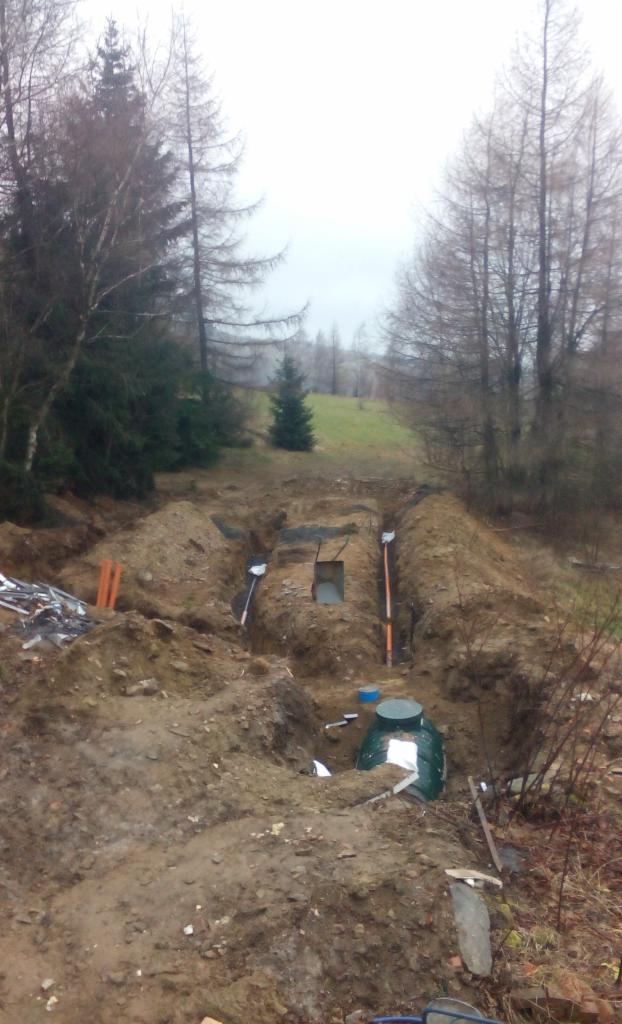 Wykop pod instalację kanalizacyjną w terenie leśnym. Widoczne rury, zbiornik, narzędzia i wykopane doły. Pochmurne niebo w tle.