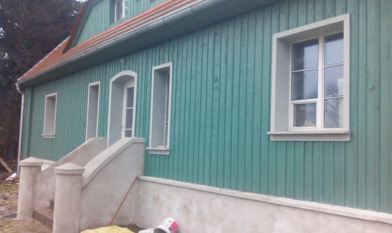 Turkusowa elewacja domu z drewnianą boazerią, białe obramowania okien i drzwi, betonowe schody wejściowe. Widoczne ślady prac wykończeniowych.