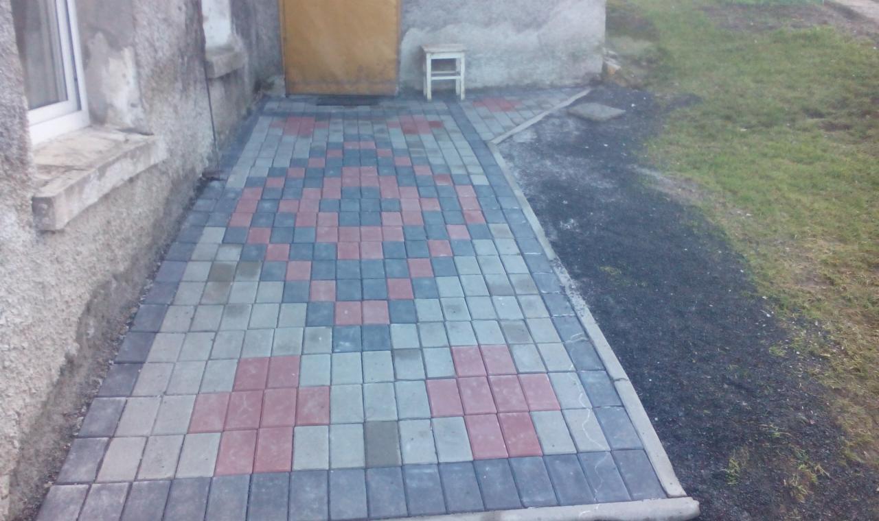 Ułożona kostka brukowa w geometryczny wzór przed budynkiem z żółtymi drzwiami i małym stołkiem. Widoczna ściana budynku i trawnik.