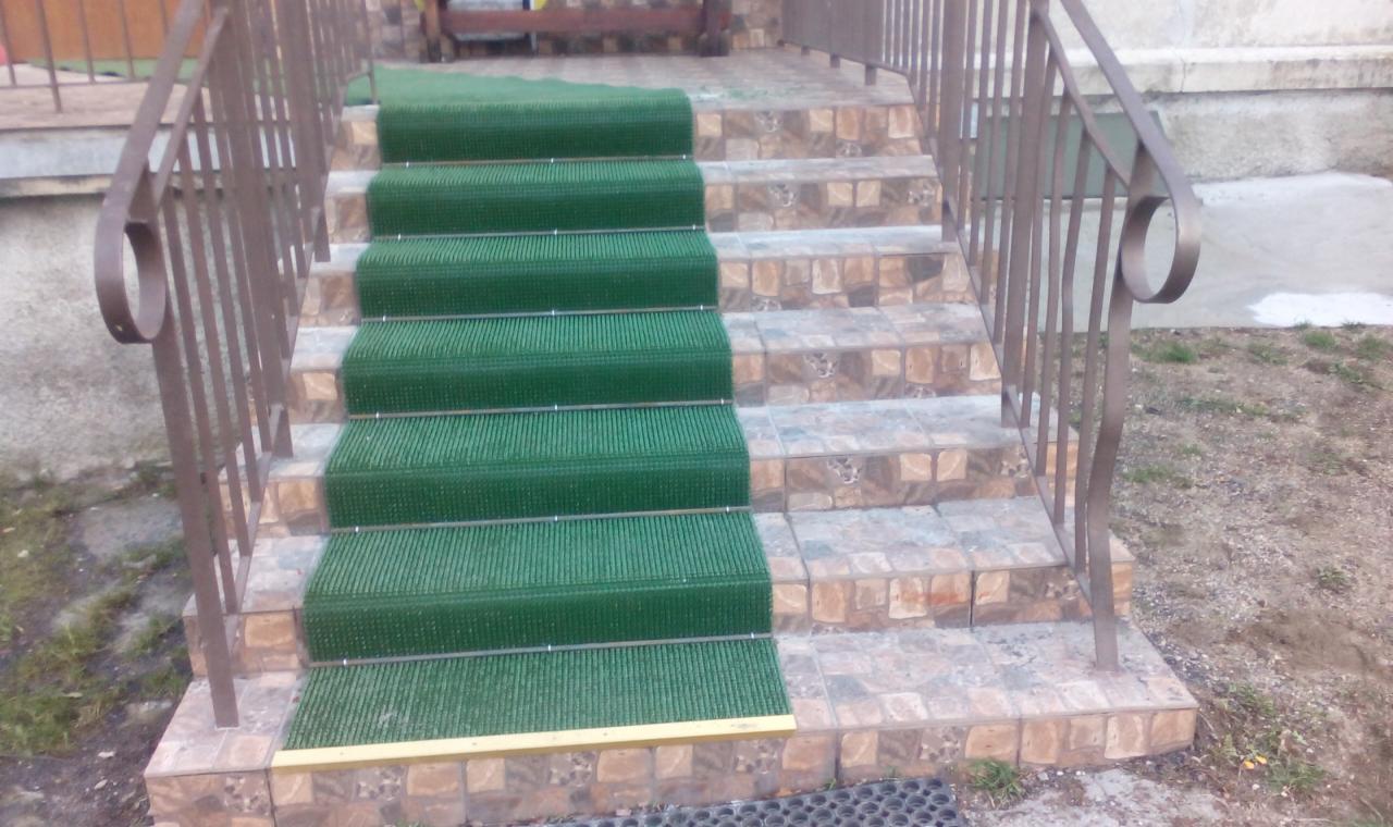 Zewnętrzne schody wyłożone płytkami z brązową balustradą i zielonymi wycieraczkami na każdym stopniu, zapewniające bezpieczeństwo i czystość.