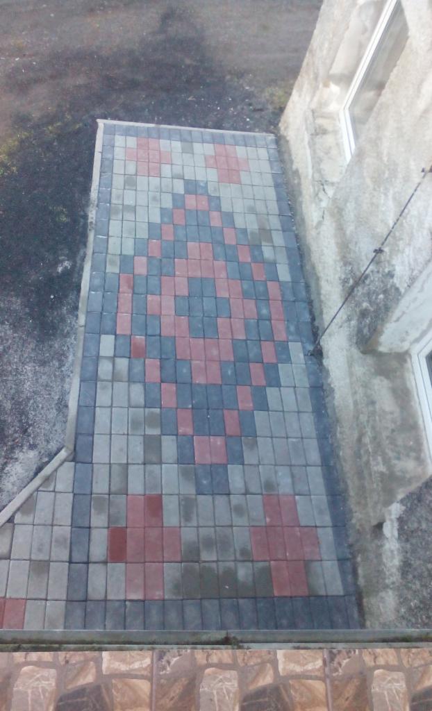 Ułożona kostka brukowa w geometryczny wzór czerwieni, szarości i granatu, tworząca chodnik przy budynku z widocznym oknem i instalacją odgromową.