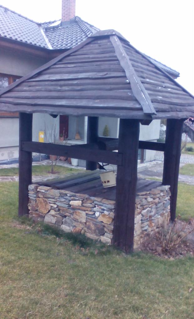 Studnia ogrodowa z kamienną obudową i drewnianą konstrukcją dachu, na tle budynku mieszkalnego. Wiadro wisi wewnątrz.