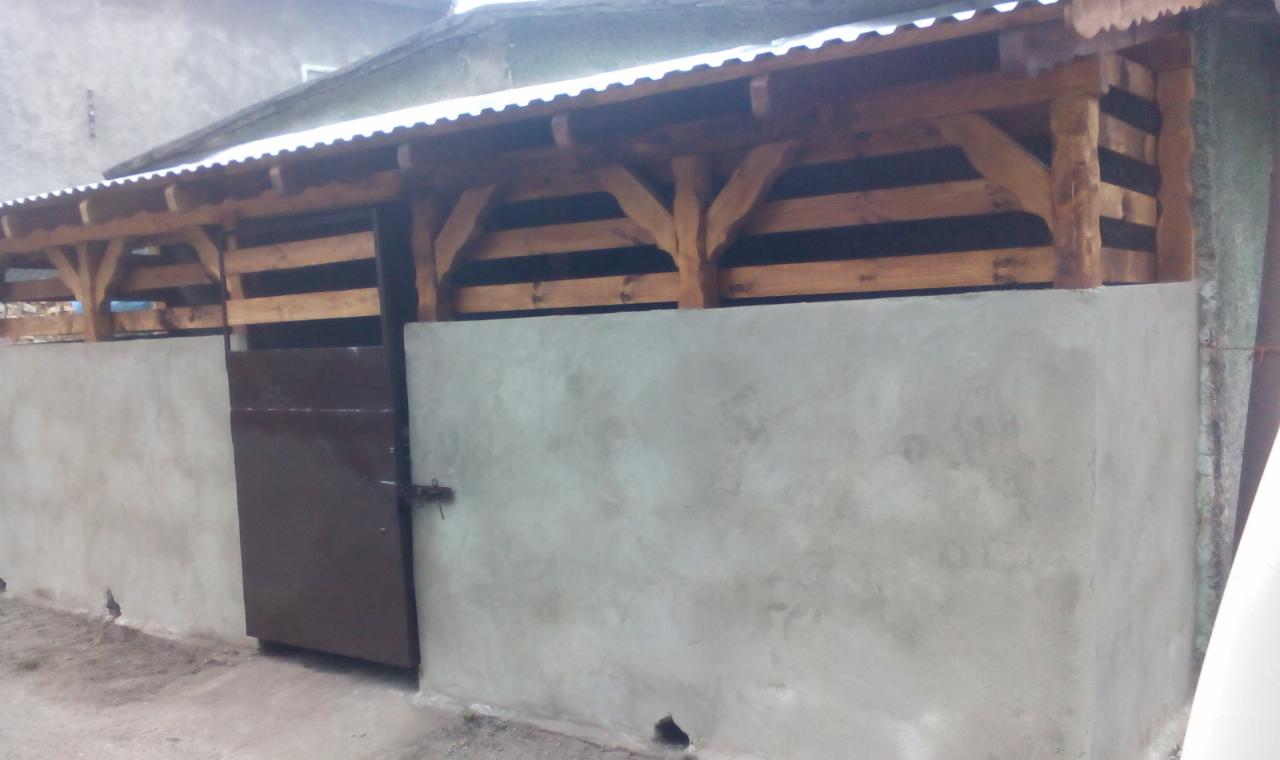 Murowana wiata z drewnianą konstrukcją dachu i metalową bramą. Ściany o surowym wykończeniu. Widok z zewnątrz.