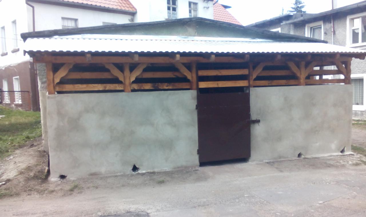 Zadaszona wiata z drewnianą konstrukcją i betonowymi ścianami, brązowe drzwi na środku. Zadaszenie z blachy falistej. Widoczne budynki w tle.