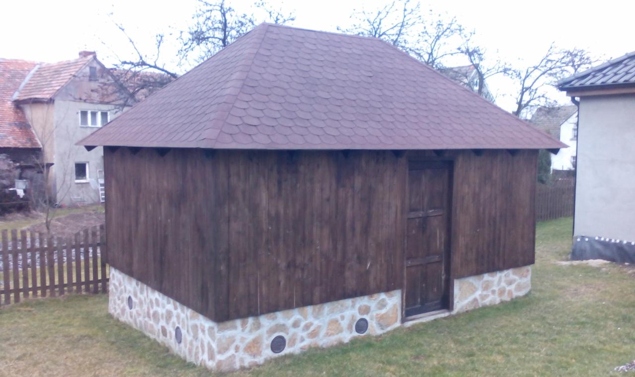 Drewniana budowla z brązowym dachem z gontów bitumicznych na kamiennej podmurówce z otworami wentylacyjnymi, otoczona trawą i budynkami w tle.