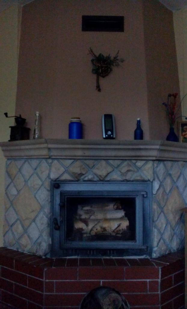 Ozdobny kominek z paleniskiem, obłożony kafelkami w romby i cegłą. Na półce dekoracje: młynek, butelki, wazon. Nad kominkiem ozdoba ścienna.