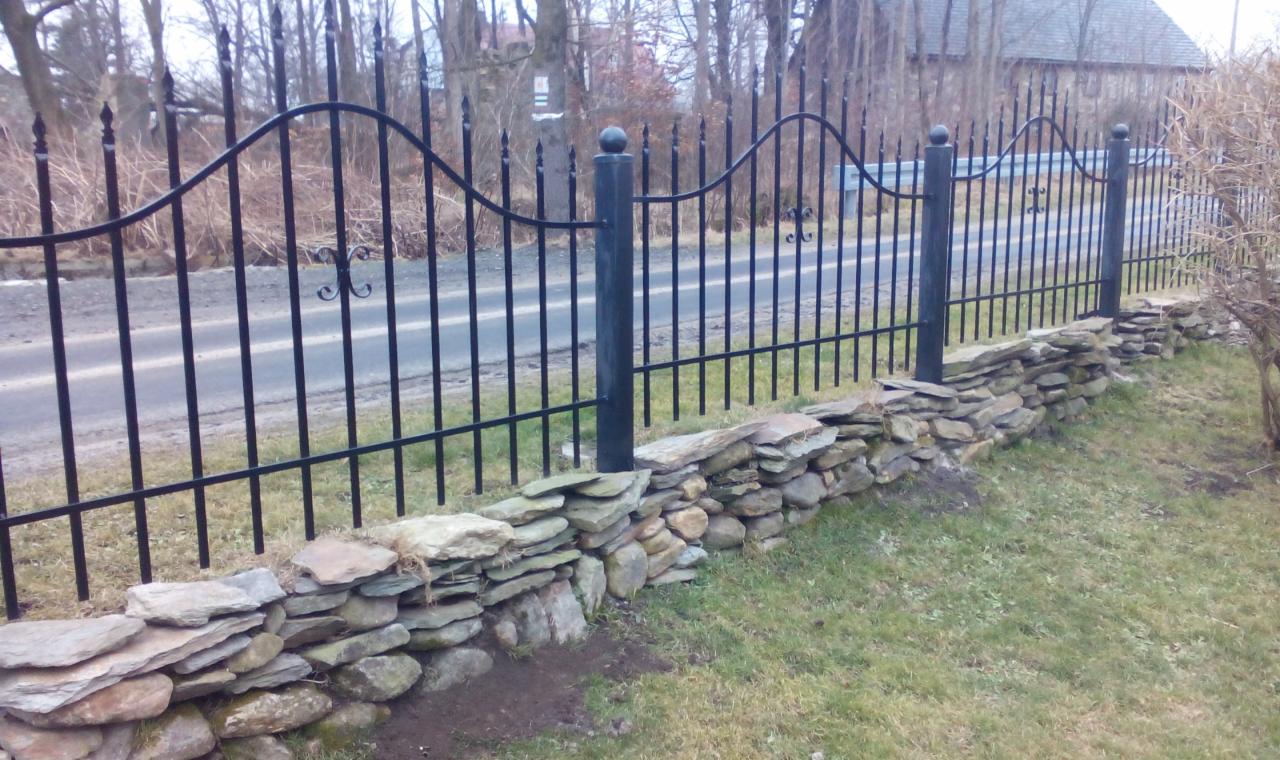 Czarny, metalowy płot z ozdobnymi elementami, osadzony na niskim murku z płaskich kamieni. W tle widoczna droga i budynek.