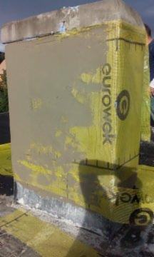 Ocieplony komin z żółtą folią Eurowek, pokryty szarą warstwą zaprawy, widoczny na tle zieleni i nieba. Detal wykończenia komina na dachu.