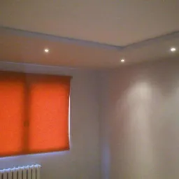 Pokój po remoncie: pomarańczowa roleta w oknie, sufit z oświetleniem punktowym i listwami dekoracyjnymi. Ściany w jasnym odcieniu. U dołu widoczny fragment grzejnika.