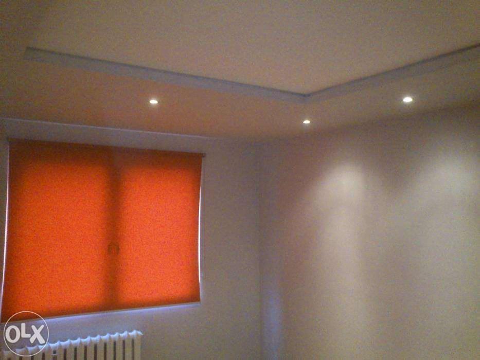 Pokój po remoncie: pomarańczowa roleta w oknie, sufit z oświetleniem punktowym i listwami dekoracyjnymi. Ściany w jasnym odcieniu. U dołu widoczny fragment grzejnika.