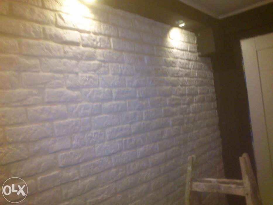 Ściana z białej cegły dekoracyjnej z oświetleniem punktowym, widoczny fragment drabiny malarskiej w prawym dolnym rogu. Tekstura cegły podkreślona przez światło.