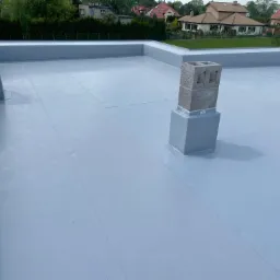 Wykończony płaski dach pokryty szarą membraną hydroizolacyjną, z dwoma kominami wentylacyjnymi, w tle domy jednorodzinne i zieleń.