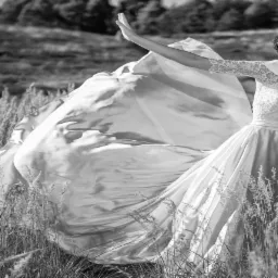 Czarno-białe zdjęcie pary młodej w polu. Suknia z koronkową górą i rozwianym dołem. Panna młoda z kwiatem we włosach, unosi rękę. Romantyczna sesja w plenerze.