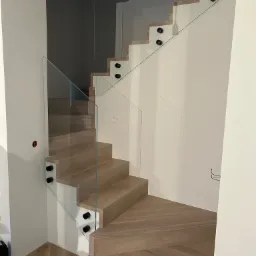 Nowoczesne schody z jasnego drewna z szklaną balustradą mocowaną na czarne punkty, widok z dołu na zakręt, parkiet w jodełkę na podłodze.