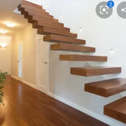 Nowoczesne, wiszące schody z drewna o ciemnym odcieniu, zamontowane przy białej ścianie z dyskretnym oświetleniem punktowym, widoczne z perspektywy podłogi z parkietem i rośliną w tle.