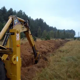 Żółta koparka CAT tworzy długi wykop w ziemi na tle lasu i pochmurnego nieba. Widoczny fragment opony i ramię maszyny w akcji.