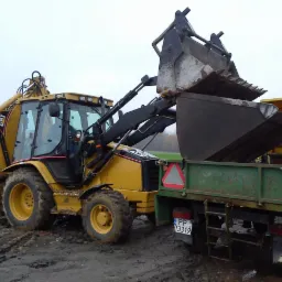 Żółta koparko-ładowarka CAT załadowuje ziemię do zielonej przyczepy na błotnistym terenie. Widoczny numer rejestracyjny przyczepy i fragment pola w tle.