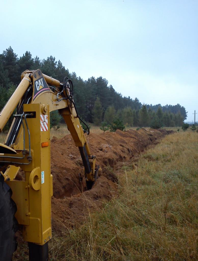 Żółta koparka CAT tworzy długi wykop w ziemi na tle lasu i pochmurnego nieba. Widoczny fragment opony i ramię maszyny w akcji.
