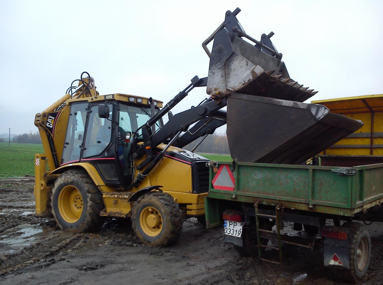 Żółta koparko-ładowarka CAT załadowuje ziemię do zielonej przyczepy na błotnistym terenie. Widoczny numer rejestracyjny przyczepy i fragment pola w tle.