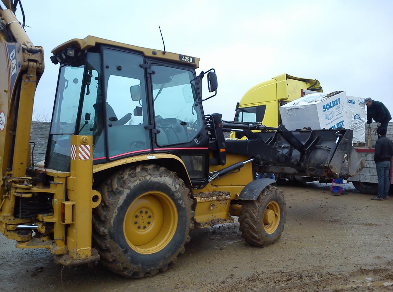 Żółta koparko-ładowarka CAT 428D na błotnistym terenie, obok ciężarówka z materiałami budowlanymi Solbet. Widoczni pracownicy nadzorujący załadunek.